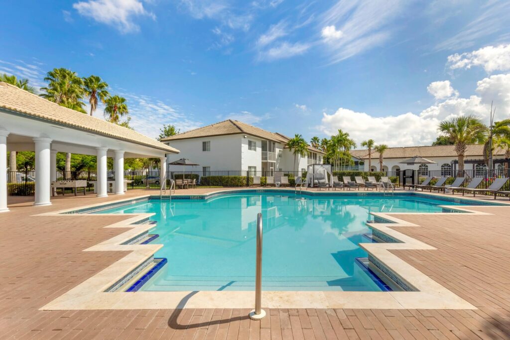 Gated pool area with lounge chairs in the back and tables with umbrella and chairs. Also covered seating area